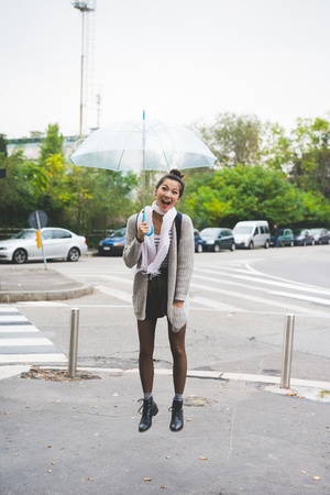 Young handsome asiatic brown hair woman jumping on the spot, holding an umbrella, looking in camera laughing - jump, rainy, having fun conceptの写真素材