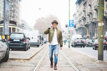 Young handsome cauciasain curly hair man walking in the street of the city, talking smartphone, overlooking pensive - communicaiton, technology, thoughtful conceptの写真素材