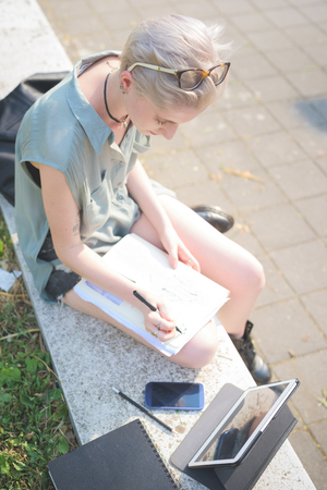 young handsome caucasian blonde italian designer sitting on a small wall, using smartphone and tablet and sketching on a notebook - creativity, enlightenment, working conceptの写真素材