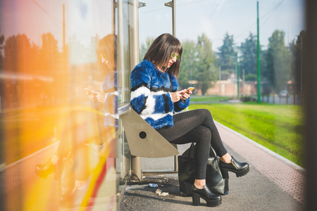 Young beautiful eastern young woman sitting at the bus stop, holding a smartphone, looking down and tapping the screen - transport, technology, social network conceptの写真素材