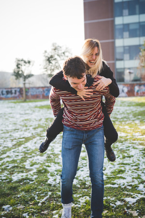 Couple of young lovers embracing outdoor in the city. She is riding piggyback, overlooking right laughing. He is looking downward, smiling - happiness, love, relationship conceptの写真素材