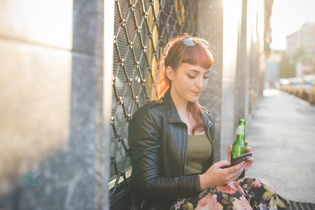 Young handsome caucasian redhead woman using smartphone holding a beer and using a smartphone, looking down and tapping the screen - social network, technology, communication concept  backlightの写真素材