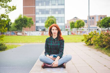 Young beautiful redhead woman sitting leg crossed in the city, holding a smartphone, looking in...