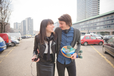 Knee figure of young caucasian woman and man with a skate and mustache walking trough the streets of the city listening music by technological device - carefreeness, friendship, love, youth conceptの写真素材
