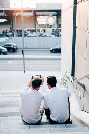 Rear view of two young modern businessman, sitting on a staircase, using a tablet, taking a selfie - technology, vanity, social network conceptの写真素材