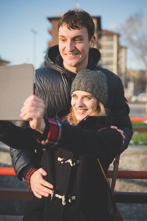 A couple of young lovers seated on sidewalk, having fun while using tablet connected internet city. She wearing brown hat, black winter coat and black jeans. He is wearing blue jeans and blue jacket.の写真素材