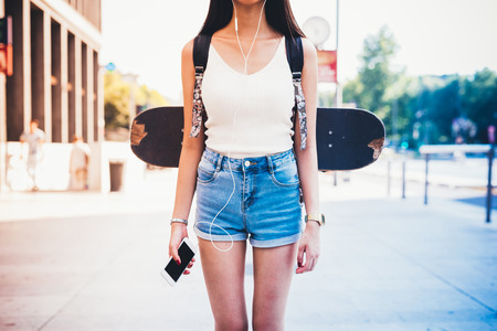 From the neck down view of young woman with skateboard and backpack, holding a smart phone and listening music with earphones - technology, sportive, music conceptの写真素材