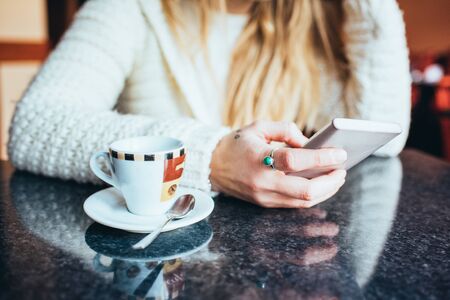Close up on the hand of a young woman sitting on a bar having a coffee, smart phone hand hold, tapping the screen - leisure, technology, social network conceptの写真素材