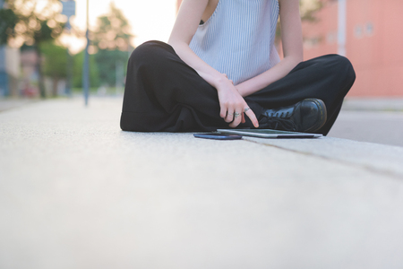 From the neck down view of young beautiful woman sitting on a sidewalk cross legged, using technological devices like smart phone and tablet, tapping the screen - technology, communication, social network conceptの写真素材