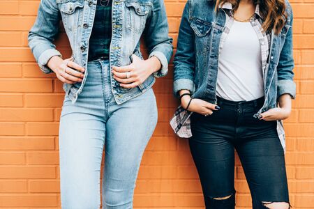 From the neck down view of two fashionable women leaning on a brick wall, wearing jeans clothes,...