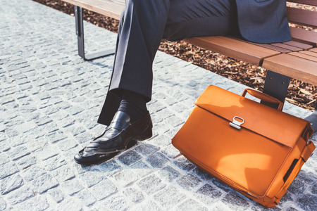 Close up on legs of contemporary businessman sitting on a bench with briefcase leaning on near him - business, work conceptの写真素材
