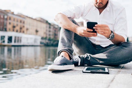Young beautiful caucasian man, sitting on the riverbank, using smart phone hand hold - technology,...
