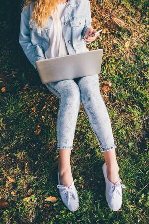 From the neck down view of young blonde woman sitting ont he grass using notebook and smart phone hand hold - technology, multitasking, social network conceptの写真素材