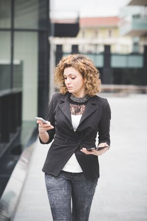 Knee figure of a young beautiful caucasian contemporary businesswoman walking through the city using smartphone and tablet looking the screen- technology, network, business, finance conceptsの写真素材