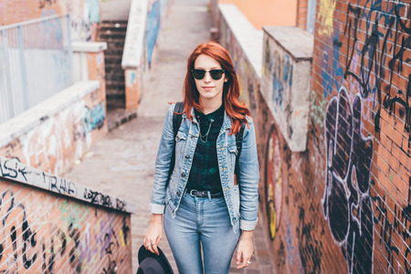 Knee figure of young handsome caucasian redhead straight hair woman posing on a staircase in the city, wearing sunglasses, looking in camera pensive - serious, thoughtful conceptの写真素材