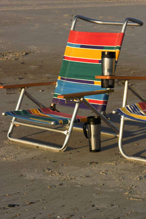 Two beach chairs along the shore with travel coffee cups. Lounge chairs set up in the sand at the beach with beverage cups. の写真素材