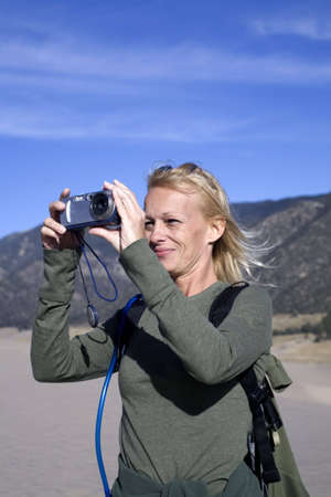 A woman hiker standing on a sand dune taking a photograph with a small cameraの写真素材