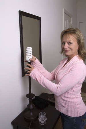 A happy woman switching out lightbulb for a more economical fluorescent bulb. Saving money by replacing bulbs at home. の写真素材