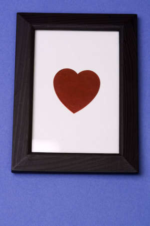 A heart symbol on a white background in a wooden black frame on a blue background.  Shot as though looking up at it on a wall. Heart is easily isolated and removed so that any text or image can be framed.の写真素材