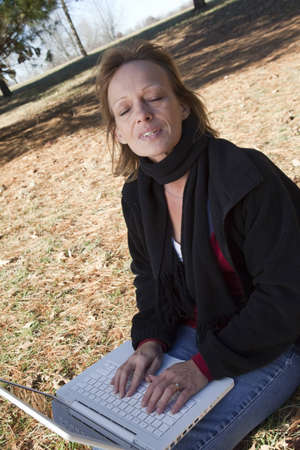 Adult woman sitting in a field daydreaming while typing on the computerの写真素材