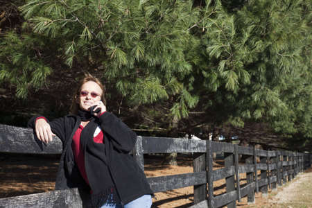 Woman talking on the phone while leaning against a fence in a rural setting.の写真素材