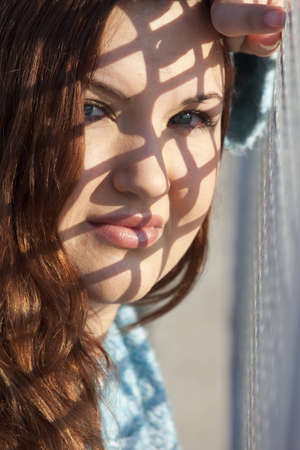 A woman leaning against a checkered fence with morning sunlight creating a pattern of shadow on her face.の写真素材