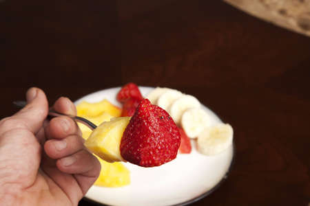 a strawberry and pineapple slice on a fork with a plate of fruit in the background.の写真素材