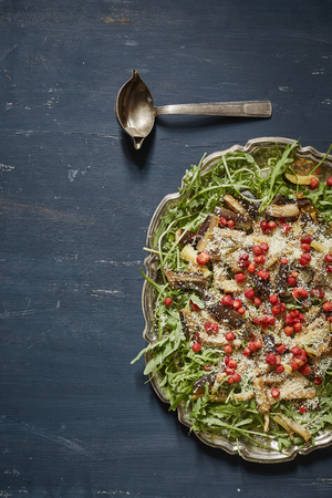 a vertical image of tasty salad with eggplant wedges, rucola, wild strawberries and grated cheese served in a vintage silver plate in a dark blue backgroundの写真素材