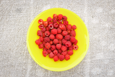 an above image of a heap of ripe raspberries on the yellow round plate in the sack background