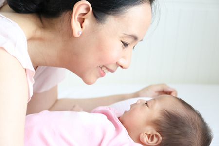 Asian mother playing with her infant baby lying on white bed backgroundの写真素材