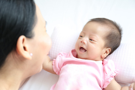 Asian mother playing with her infant baby lying on white bed backgroundの写真素材