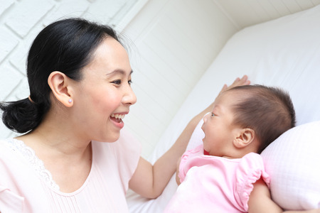 Asian mother playing with her infant baby lying on white bed backgroundの写真素材
