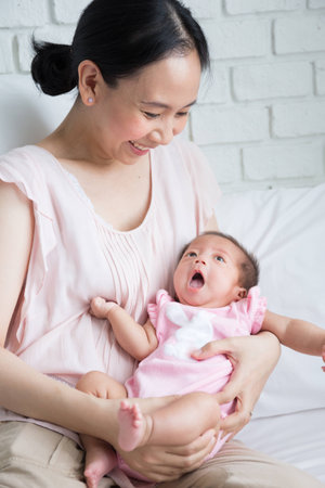 Asian mother playing with her infant baby lying on white bed backgroundの写真素材