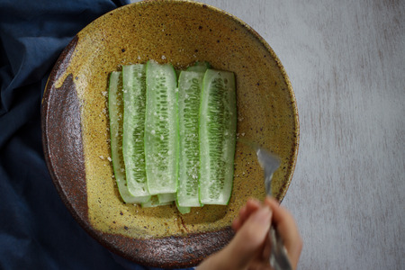 Slices of cucumber with sea salt served on old yellow plate and moving hand, still life imageの写真素材