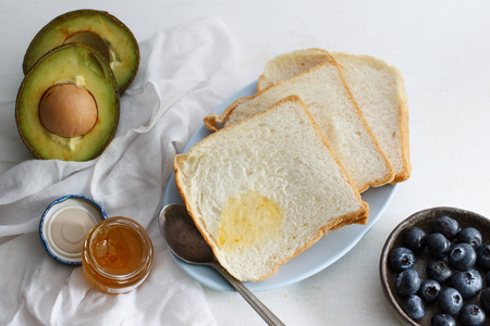 Sliced hokkaido milk bread, organic avocado, orange jam, blueberry and book put on white table.の写真素材