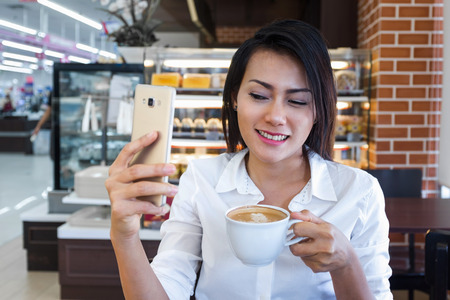 Asia women who drink coffee and look at his cell phone at the same time in a cafe, focus on the face.の写真素材