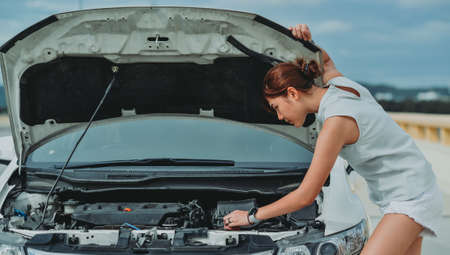 Young woman standing near broken down car with popped up hood having trouble with her vehicle. Female driver waiting for help beside malfunction auto.の写真素材