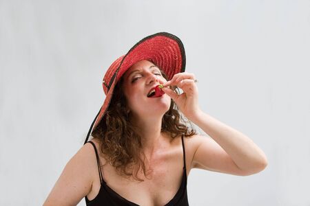 Beautiful middle aged woman wearing a red hat and biting on a strawberry, isolated