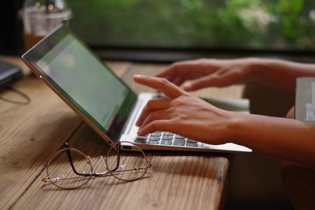 Woman's hand working on keyboard laptop computer on wood table in a cafe ,Blurred and soft focus imageの写真素材