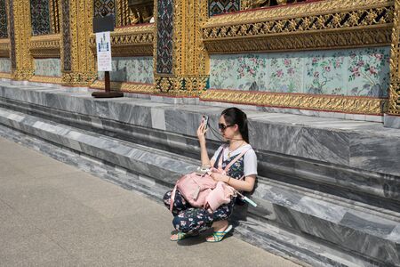 Bangkok, Thailand - October 15, 2018: Chinese female tourists leaning against the wall of temple of Emerald Buddha for taking a photo with smartphone at Wat Phra Kaew, Bangkokのeditorial素材