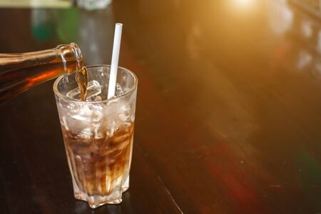sparkling water soft drinks are pouring from bottle into glass with ice on wood table with sunlight...