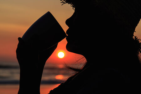 Silhouette of Thai woman drinking holding coffee cup at the beach in sunset timeの写真素材