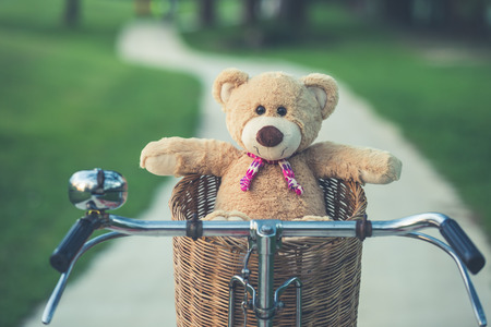 Close up lovely brown teddy bear in rattan basket on vintage bike in green field with lens flare. Warm toning effect. Retro and vintage styleの写真素材