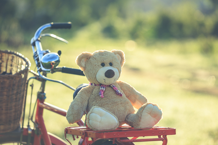 Close up lovely brown teddy bear in rattan basket on vintage bike in green field with lens flare. Warm toning effect. Retro and vintage styleの写真素材