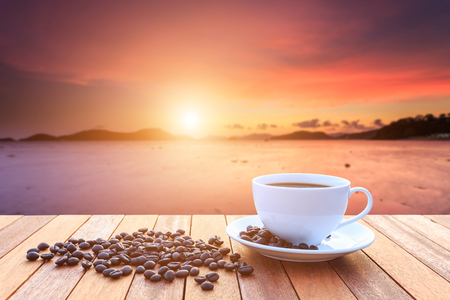 Close up white coffee cup and coffee beans on wood table and view of sunset or sunrise backgroundの写真素材