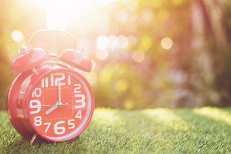 Close up red alarm clock on green grass with sunlight blur and bokeh background in morning timeの写真素材