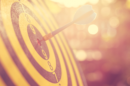 Close up blue dart arrow in the center of dartboard. Blur and bokeh in sunrise time background. Warm vintage effect styleの写真素材