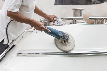 Yacht maintenance. A man polishing side of the white boat by grinder machine in the marinaの写真素材