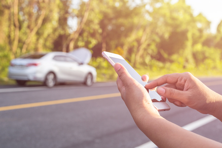 Close up hand of woman using smartphone and blur of her broken car parking on the road. Contacting car technician or need help conceptの写真素材