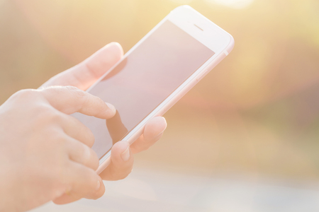 Close up hand of woman holding or using smartphone on green blur and sunlight effectの写真素材